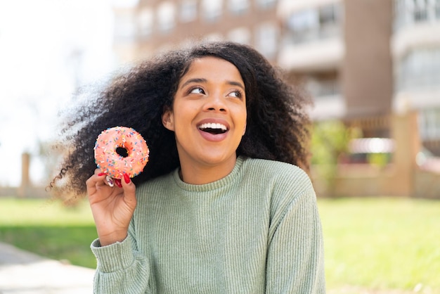Młoda Afroamerykanka trzymająca pączka na zewnątrz patrząca w górę uśmiechając się