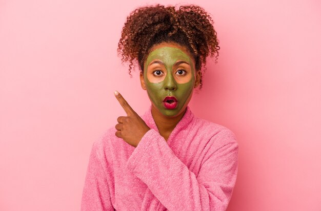 Młoda afroamerykanka ubrana w szlafrok i maskę na twarz na różowym tle, wskazującą na bok