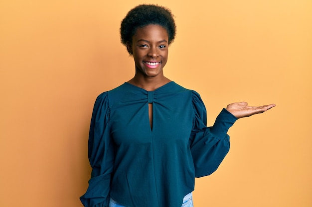 Młoda Afroamerykanka ubrana w zwykłe ubrania, uśmiechnięta wesoło, przedstawiająca i wskazująca dłonią patrząc w kamerę