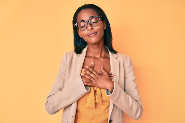Młoda Afroamerykanka w ubraniach biznesowych uśmiechnięta z rękami na klatce piersiowej z zamkniętymi oczami i wdzięcznym gestem na twarzy. koncepcja zdrowia.