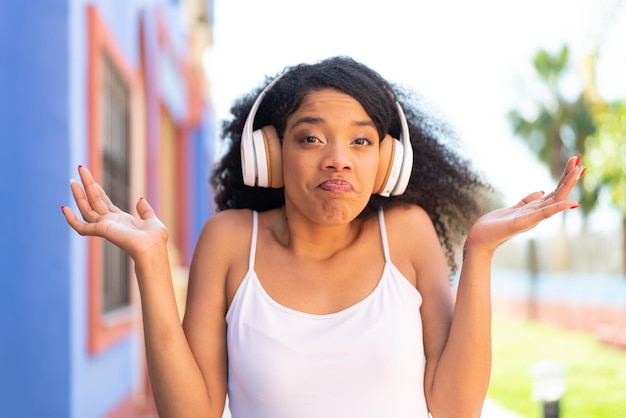 Młoda Afroamerykanka z słuchawkami na zewnątrz robiąca gest wątpliwości