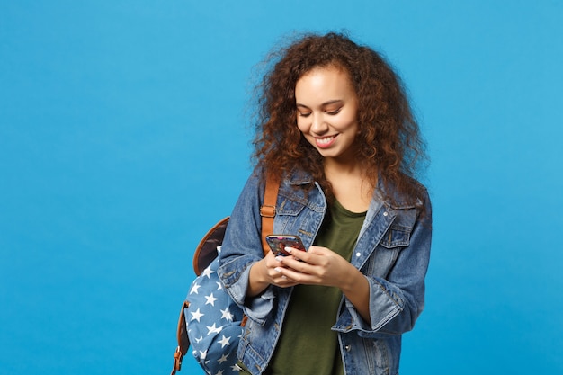 Młoda Afroamerykańska Dziewczyna Nastoletnia Studentka W Dżinsowych Ubraniach, Plecak Trzyma Telefon Na Białym Tle Na Niebieskiej ścianie