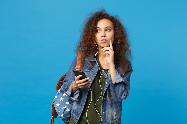 Młoda afroamerykańska dziewczyna nastoletnia studentka w dżinsowych ubraniach, słuchawki plecakowe izolowane na niebieskiej ścianie