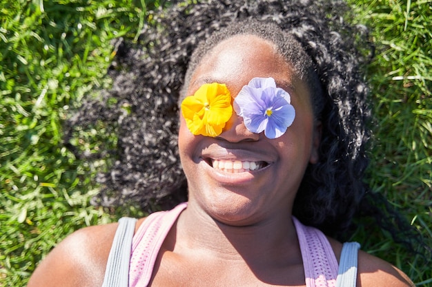 Młoda afroamerykańska kobieta leżąca na trawie uśmiecha się z dwoma kwiatami w oczach