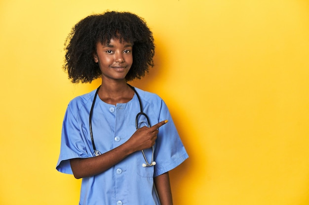 Młoda afroamerykańska pielęgniarka w studiu na żółtym tle uśmiecha się i wskazuje na bok