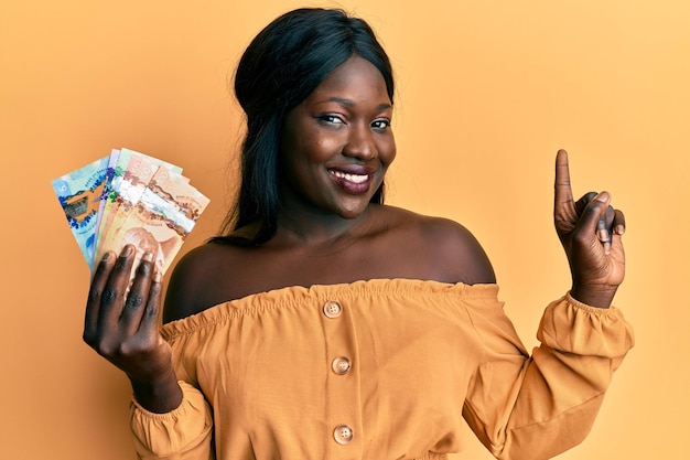 Młoda afrykańska kobieta trzymająca kanadyjskie dolary uśmiechając się szczęśliwie wskazując ręką i palcem na bok