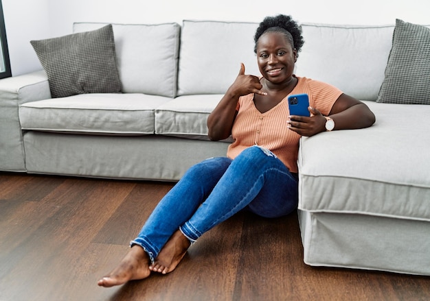 Młoda afrykańska kobieta za pomocą smartfona siedzącego na podłodze w domu uśmiecha się, wykonując gest telefoniczny ręką i palcami, jak rozmawiając przez telefon, komunikując koncepcje
