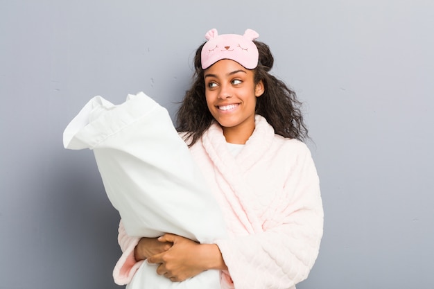 Młoda amerykanin afrykańskiego pochodzenia kobieta jest ubranym piżamę i sen maskę trzyma poduszkę ono uśmiecha się ufny z skrzyżowanymi rękami.