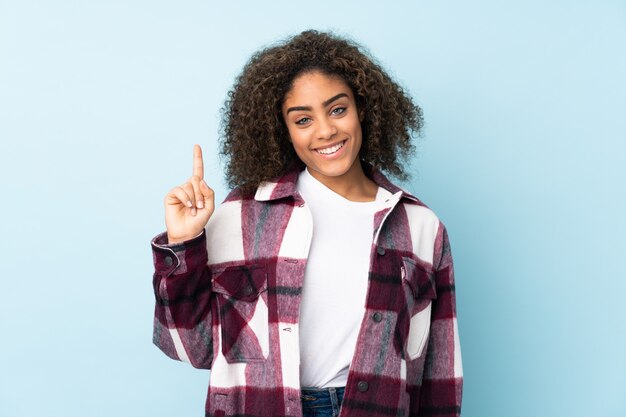 Młoda amerykanin afrykańskiego pochodzenia kobieta na błękit ścianie pokazuje i podnosi palec w znaku najlepszy