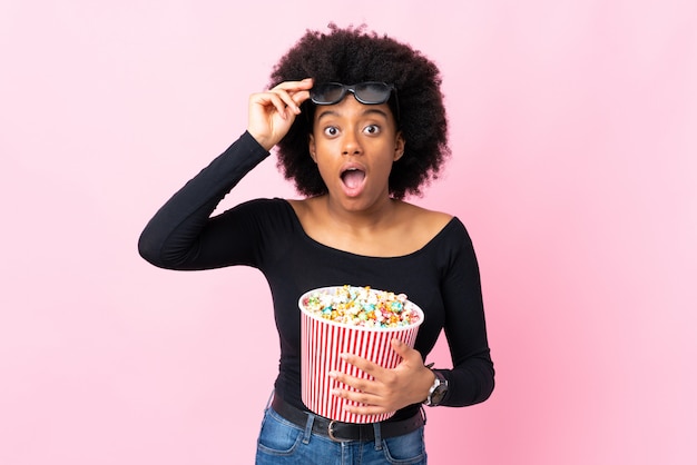 Młoda amerykanin afrykańskiego pochodzenia kobieta odizolowywająca na menchii ścianie zaskakującej z 3d szkłami i trzymający dużego wiadro popcorns