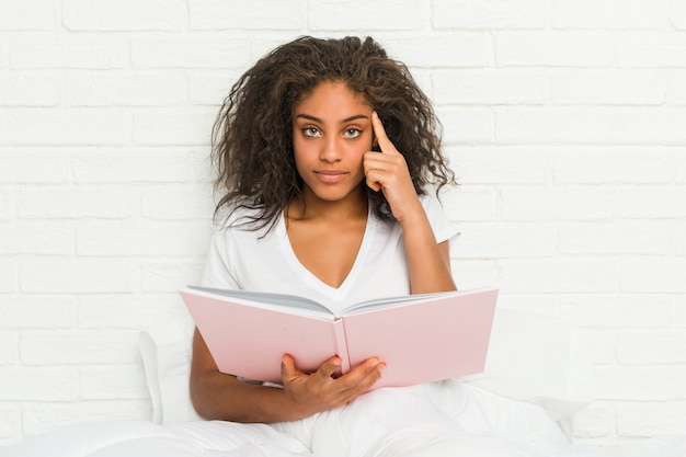 Młoda amerykanin afrykańskiego pochodzenia kobieta siedzi na łóżku studiuje wskazujący jego świątynię palcem, myśleć, skupiający się na zadaniu.