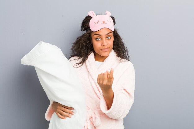 Młoda amerykanin afrykańskiego pochodzenia kobieta w piżamie i masce do spania trzyma poduszkę wskazując palcem na ciebie, jakby zapraszając podejść bliżej.