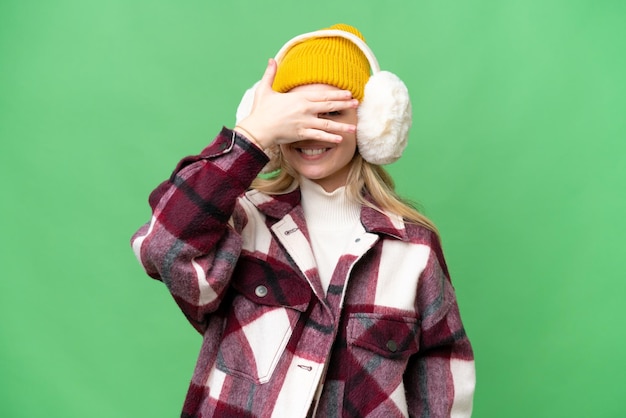 Młoda Angielka ubrana w zimowe mufki na odizolowanym tle zakrywająca oczy rękami i uśmiechająca się