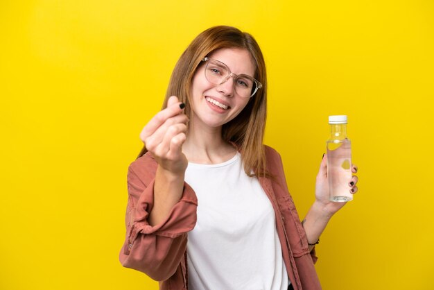 Młoda Angielka z butelką wody na żółtym tle robi pieniądze gest