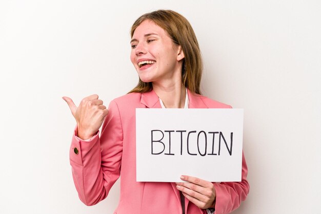 Młoda angielska kobieta biznesu trzymająca tabliczkę bitcoin na białym tle wskazuje palcem kciuka, śmiejąc się i beztrosko