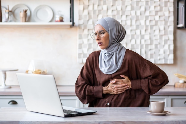 Młoda arabka w hidżabie pracuje, studiując w domu, korzystając z laptopa online, trzymając się za klatkę piersiową