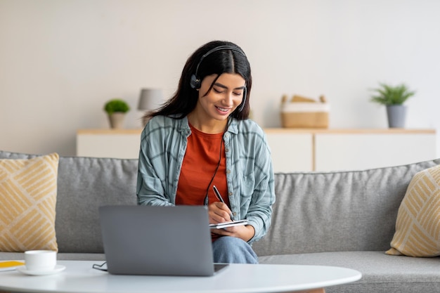 Młoda Arabka w słuchawkach, korzystająca z laptopa, pracująca lub studiująca online z domu, robiąca notatki podczas spotkania biznesowego