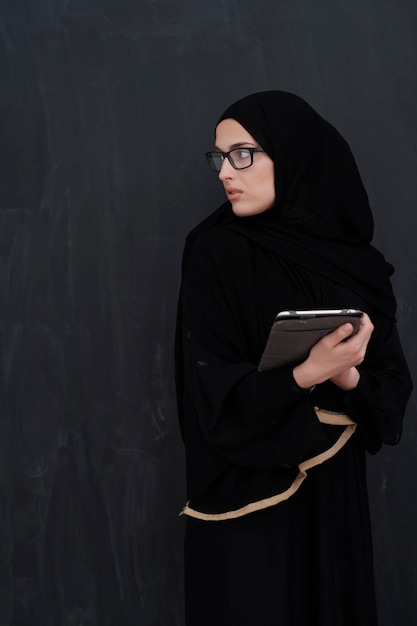 Młoda arabska bizneswoman w tradycyjnych strojach lub abaya i okularach trzymająca komputer typu tablet przed czarną tablicą reprezentującą nowoczesną modę i technologię islamu