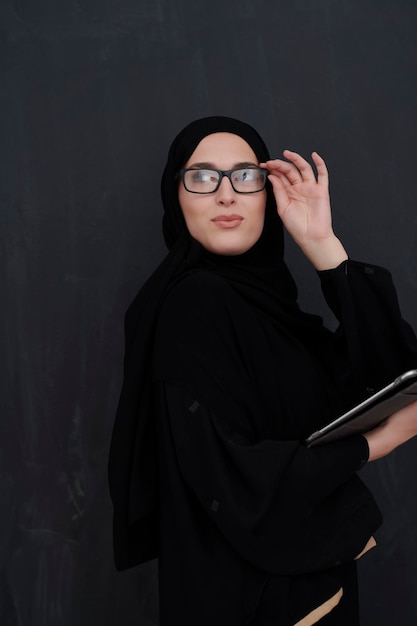 Młoda arabska bizneswoman w tradycyjnych strojach lub abaya i okularach trzymająca komputer typu tablet przed czarną tablicą reprezentującą nowoczesną modę i technologię islamu