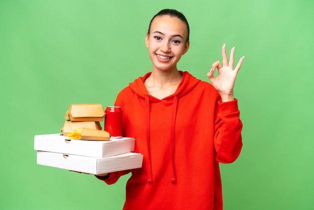 Młoda arabska kobieta trzymająca fast food na odosobnionym tle pokazująca znak ok palcami