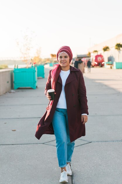 Młoda arabska muzułmanka w hidżabie spaceruje po ulicy nadmorskiej i pije kawę na wynos