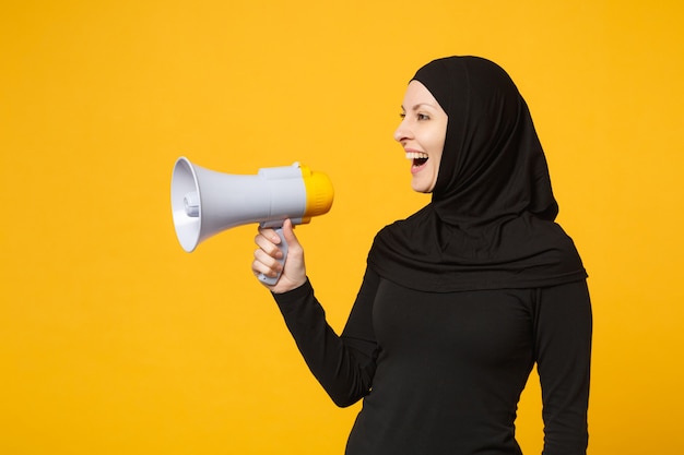 Młoda Arabska Muzułmańska Kobieta W Hidżab Czarne Ubrania Trzyma W Ręku Bullhorn Adres Publiczny Megafon Na Białym Tle Na Portret żółtej ściany. Koncepcja życia Religijnego Ludzi.