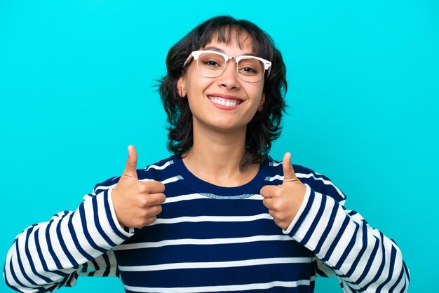 Młoda argentyńska kobieta na białym tle na niebieskim tle w okularach i z kciukiem do góry
