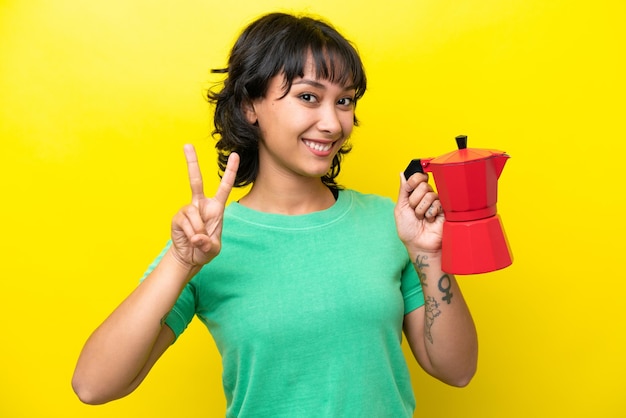Młoda argentyńska kobieta trzymająca dzbanek do kawy na żółtym tle uśmiechnięta i pokazująca znak zwycięstwa