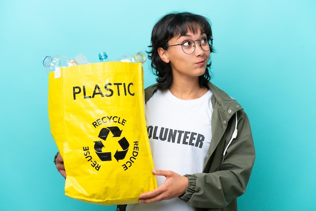 Młoda argentyńska kobieta trzymająca torbę pełną plastikowych butelek do recyklingu na białym tle na niebieskim tle i patrząc w górę