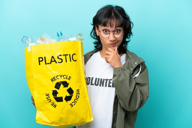 Młoda argentyńska kobieta trzymająca torbę pełną plastikowych butelek do recyklingu na białym tle na niebieskim tle, mająca wątpliwości i myśląca