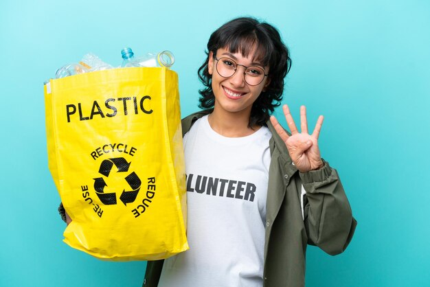 Młoda argentyńska kobieta trzymająca torbę pełną plastikowych butelek do recyklingu odizolowana na niebieskim tle szczęśliwa i licząca cztery palcami