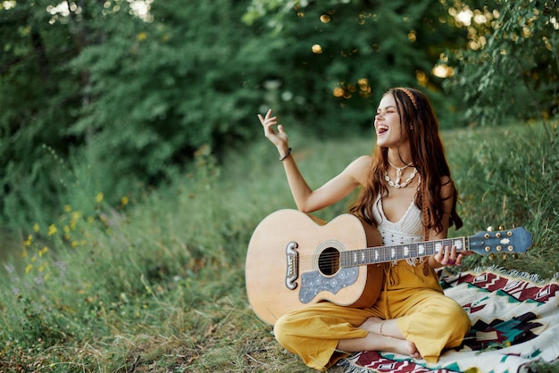 Młoda artystka hipiska gra na gitarze i śpiewa piosenki w ekologicznych ubraniach, siedząc na ziemi na zewnątrz jesienią, patrząc na zachód słońca