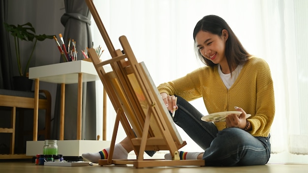 Młoda artystka w żółtym swetrze z paletą w dłoni maluje na płótnie na podłodze.