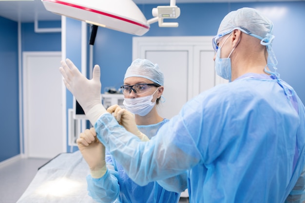 Młoda asystentka w mundurze pomaga chirurgowi założyć rękawiczki medyczne przed operacją w szpitalu