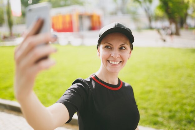 Młoda Atletyczna Uśmiechnięta Piękna Brunetka Dziewczyna W Czarnym Mundurze, Czapka Patrząc Na Smartfonie I Robi Selfie Na Telefonie Komórkowym Podczas Treningu W Parku Miejskim Na Zewnątrz