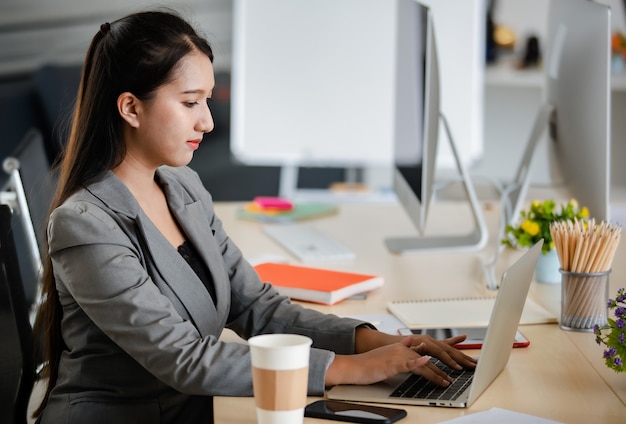 Młoda atrakcyjna Azjatycka kobieta w szarym garniturze pracuje na laptopie w nowocześnie wyglądającym biurze. Koncepcja nowoczesnego stylu życia w biurze.