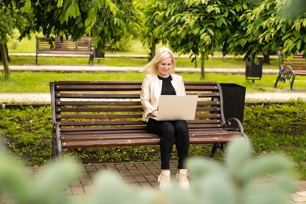 Młoda atrakcyjna bizneswoman za pomocą komputera przenośnego pc siedząc na drewnianej ławce w parku miejskim.