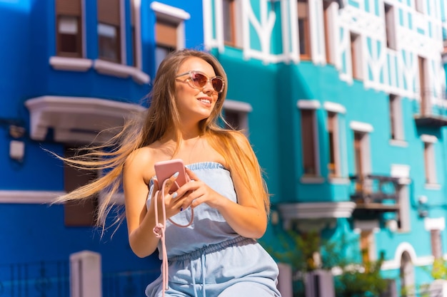 Młoda atrakcyjna blondynka patrząca na telefon komórkowy i jej włosy wiejące wiatrem za kolorową żółtą i zieloną fasadą