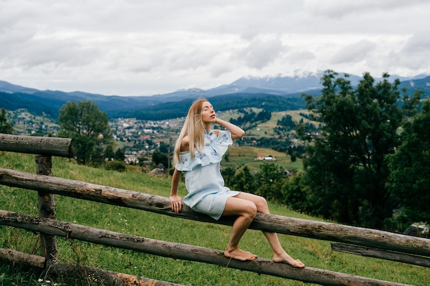 Młoda atrakcyjna elegancka blondynka w niebieskiej sukience siedzi na drewnianym płocie na wsi