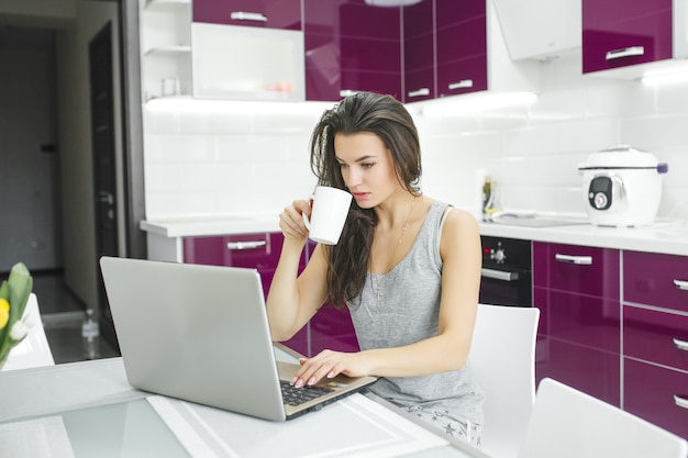 Młoda atrakcyjna kobieta pracuje na komputerze osobistym przy kuchnią. Kobieta pracuje przy komputerze w godzinach porannych. Freelancer przy laptopie kryty.