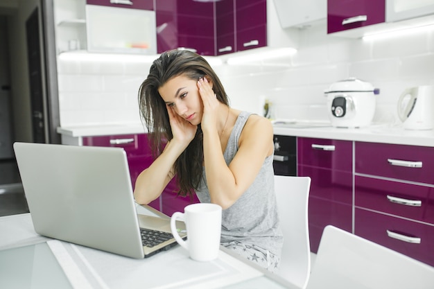 Młoda atrakcyjna kobieta pracuje na laptopie w kuchni. Kobieta pisze na komputerze w pomieszczeniu rano.
