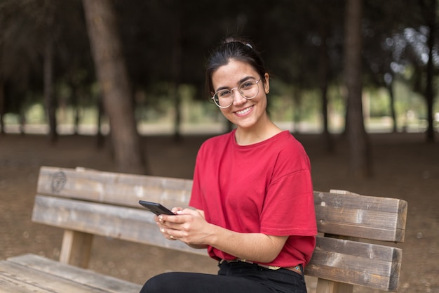 Młoda atrakcyjna kobieta siedzi na ławce i używa jej telefon w parku z szkłami, z czerwoną koszula. Sewilla, Hiszpania