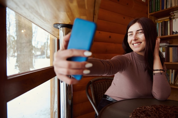 Młoda Atrakcyjna Kobieta Trzyma Smartfona I Uśmiecha Się, Robiąc Selfie, Siedząc W Kawiarni Na Tle Drewnianej ściany I Półek Z Książkami