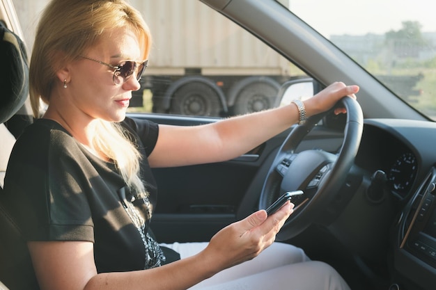 Młoda atrakcyjna kobieta w okularach przeciwsłonecznych siedzi za kierownicą samochodu ze smartfonem w dłoni