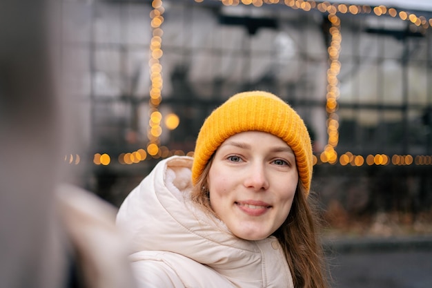 Młoda atrakcyjna kobieta w żółtym kapeluszu robi sobie selfie na tle girlandy na ulicy zimą Obraz do Twojego projektu