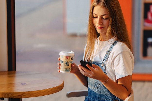 Młoda atrakcyjna ładna dziewczyna przy kawiarnią z kawą i telefonem przy rankiem promieniuje