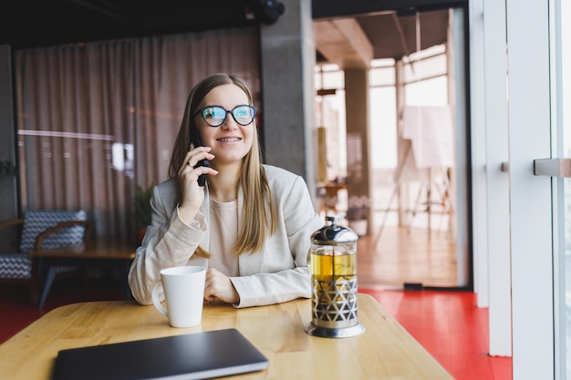 Młoda atrakcyjna menedżerka rozmawia przez telefon komórkowy i uśmiecha się, siedząc samotnie przy dużym oknie w kawiarni w wolnym czasie i pracując na komputerze Szczęśliwa kobieta odpoczywa w kawiarni