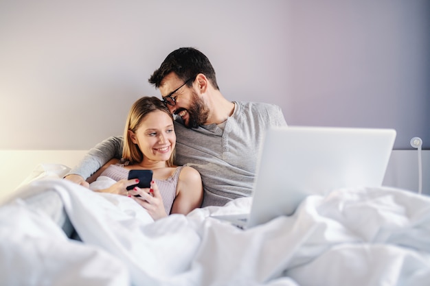 Młoda atrakcyjna para siedzi na łóżku w sypialni i przytulanie. Obsługuje mieć laptop na kolanach podczas gdy kobieta trzyma mądrze telefon.