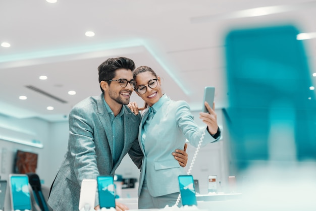 Młoda atrakcyjna para wielokulturowa ubrana elegancko biorąc selfie z nowym smartfonem w sklepie tech.