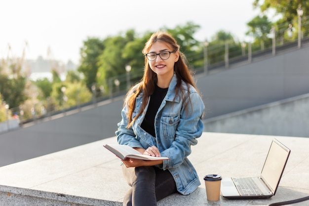 Młoda atrakcyjna studencka kobieta czyta książkę podczas gdy siedzący na schodkach w mieście. Pojęcie kształcenia na odległość. Współczesny student